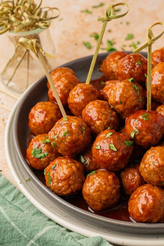 Plate of glazed meatballs garnished with herbs, some skewered with bamboo picks, set beside a jar of skewers and green napkin.