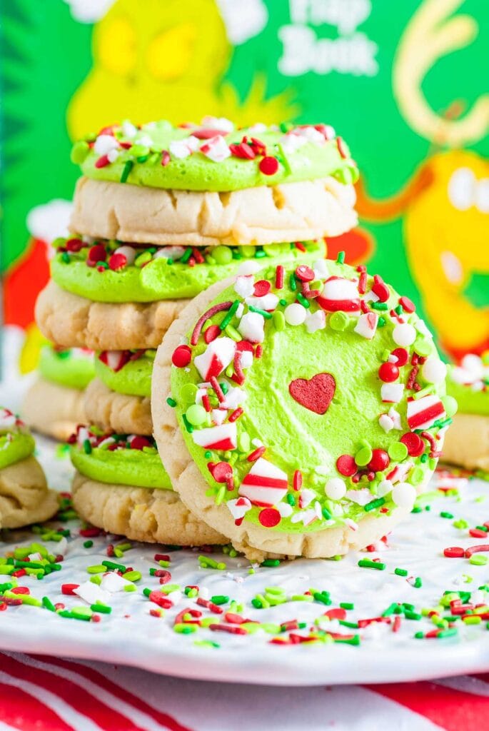 Stacked sugar cookies with green frosting, red heart, and festive sprinkles, arranged on white plate with Grinch and Max illustration in background.