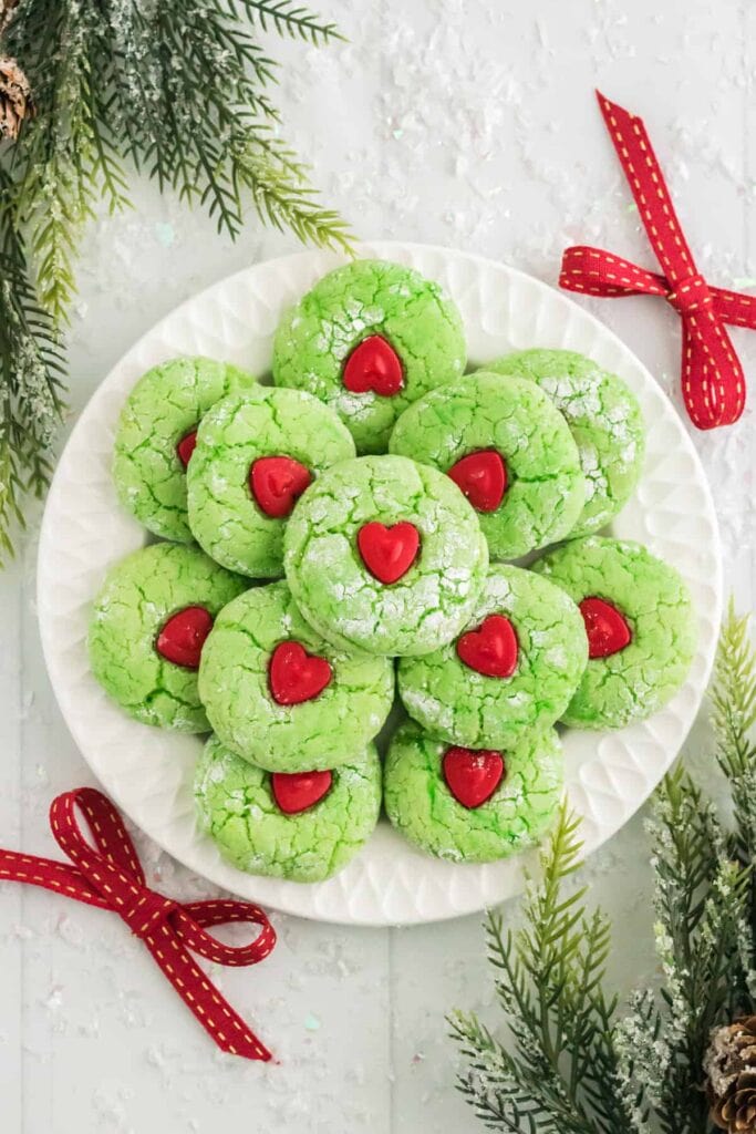 Bright green cracked cookies dusted with powdered sugar, each topped with red heart candy, arranged on white plate surrounded by festive evergreens and red ribbons.