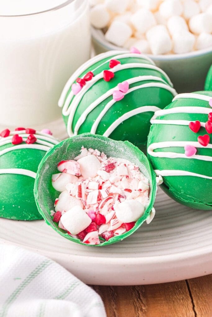 Green chocolate cocoa bombs with white drizzle and heart sprinkles, one cut open showing marshmallows and peppermint, set beside milk and marshmallow bowl.