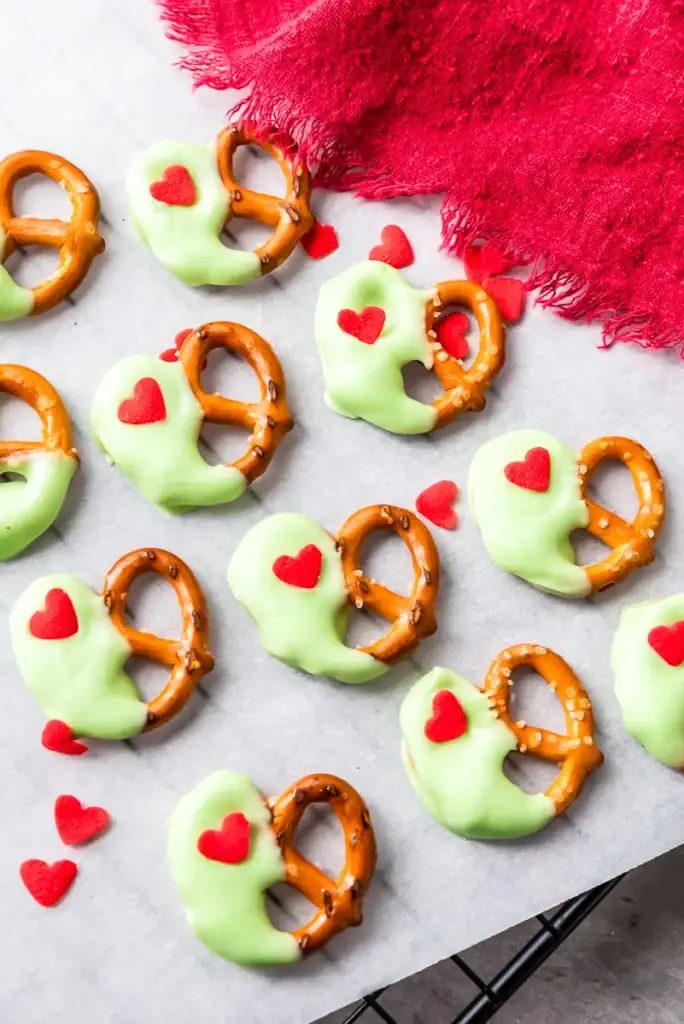 Small pretzels dipped in light green coating with red heart decorations, arranged on parchment with festive red cloth and scattered hearts.