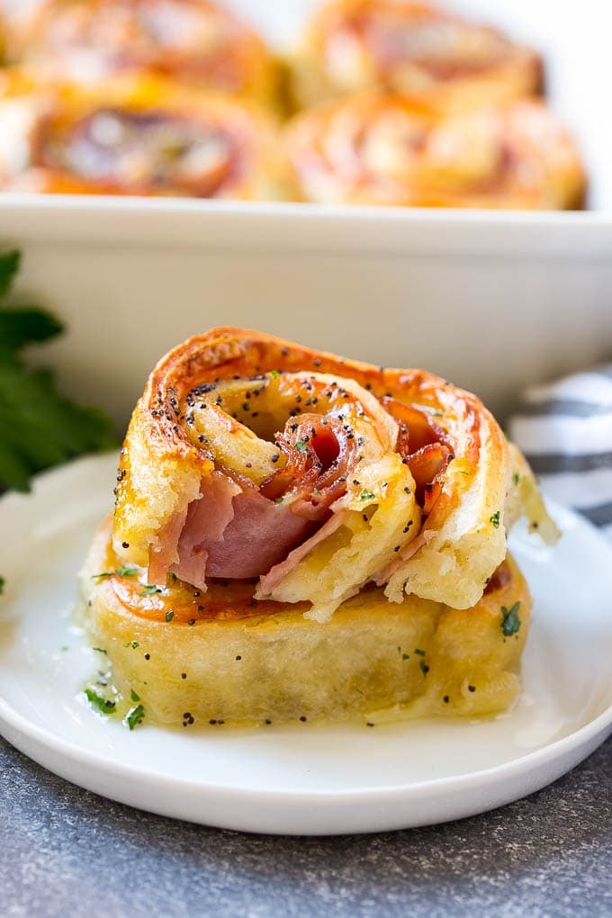 Golden flaky roll filled with ham and melted cheese, topped with poppy seeds and herbs, served on a white plate with baking dish in background.