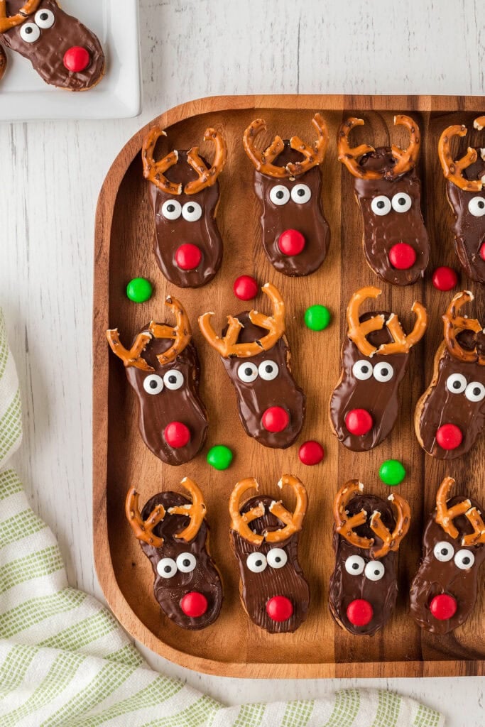 Chocolate-coated oval cookies decorated as reindeer faces with candy eyes, red noses, and pretzel antlers, arranged on wooden tray with scattered candies and striped cloth.