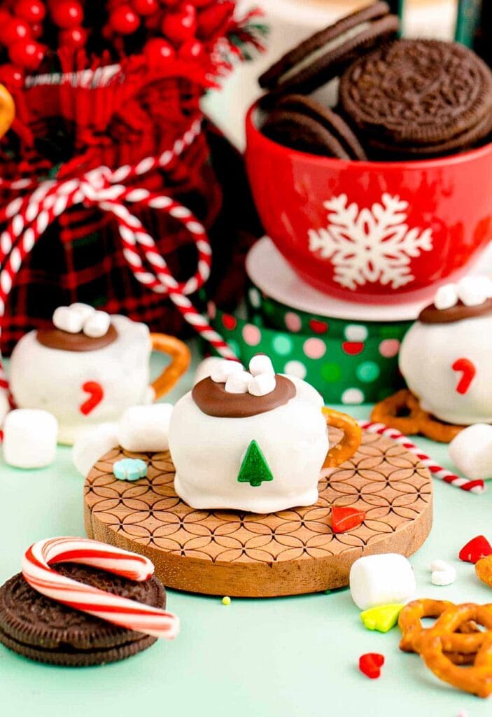 Mini mug-shaped desserts with pretzel handles, chocolate tops, marshmallows, and festive decorations, arranged with holiday candies and ribbons.