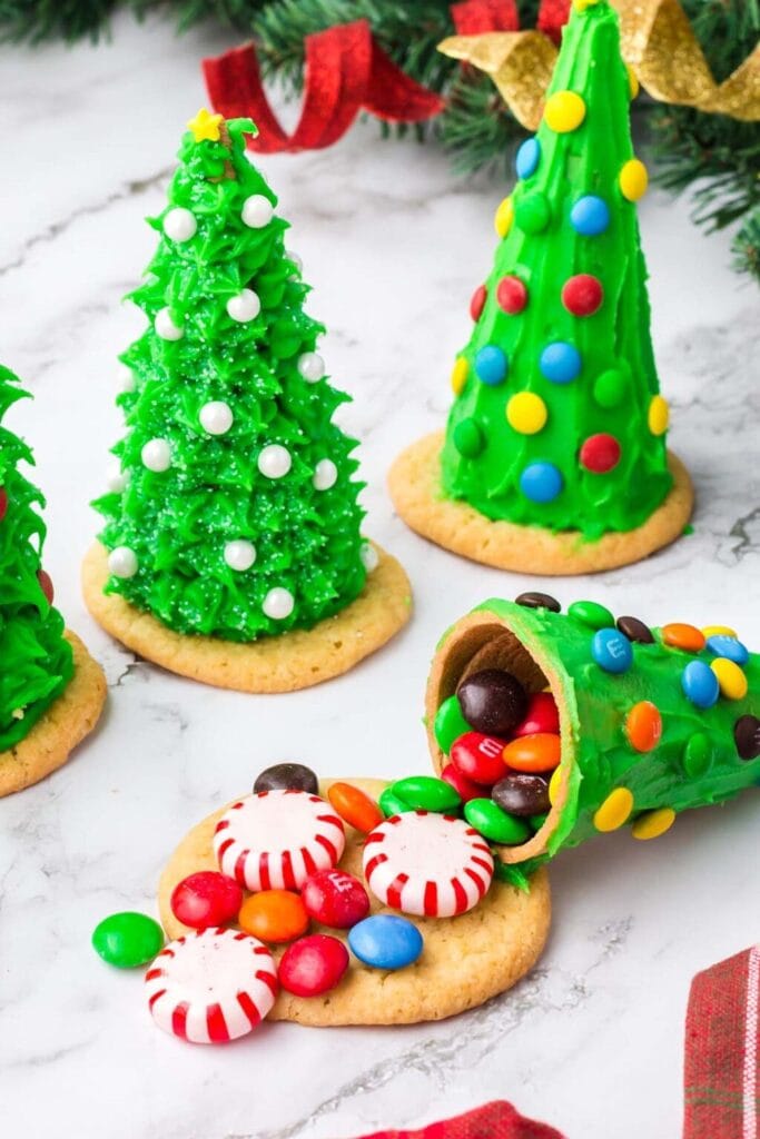 Edible Christmas trees made from green frosted cones decorated with candies and sprinkles, one tipped to reveal peppermint and M&M filling, set among pine branches and holiday ribbons.