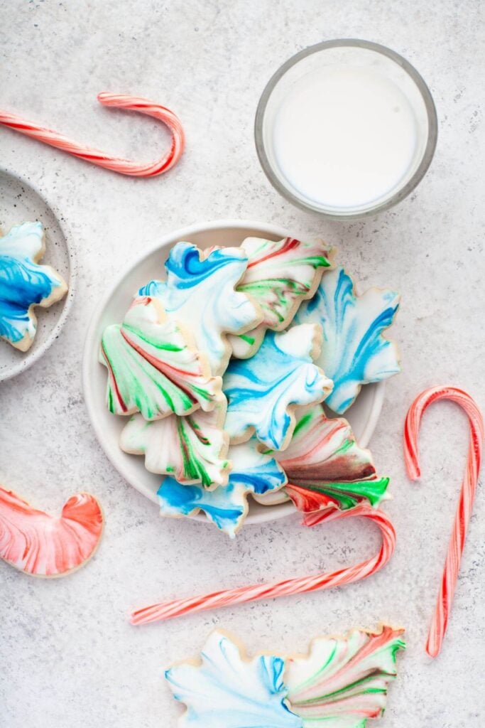 Leaf-shaped sugar cookies with colorful marbled icing in red, blue, green, and white, arranged on plate with candy canes and glass of milk.
