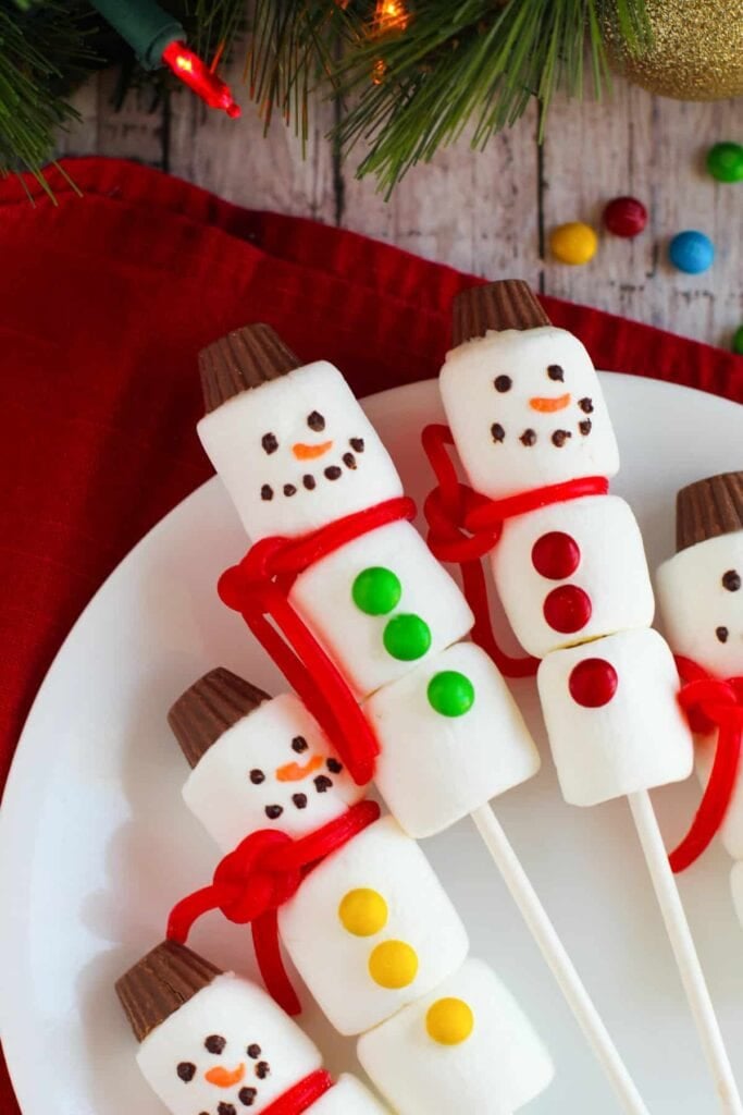 Snowman marshmallow pops with peanut butter cup hats, candy faces, colorful buttons, and red licorice scarves, arranged on plate with festive cloth and Christmas tree decor.