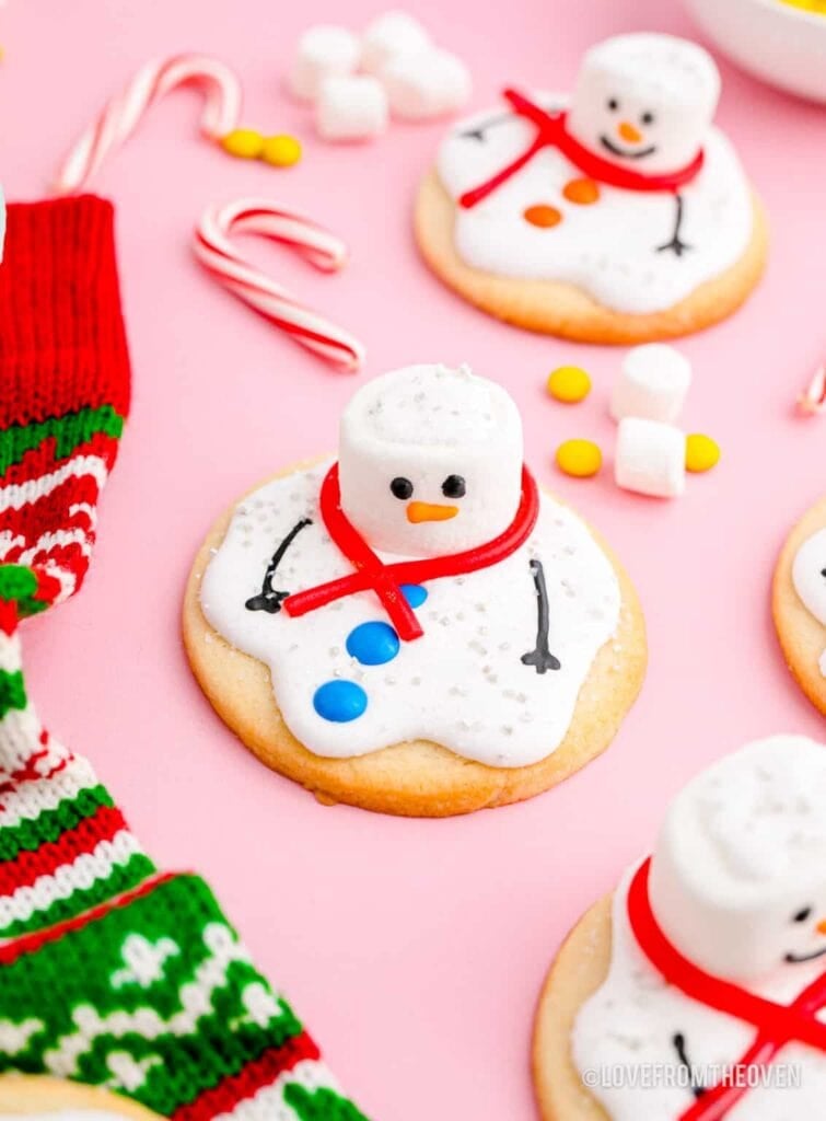 Round cookies topped with marshmallow snowmen on melted icing, decorated with candy scarves, buttons, and arms, arranged on pink surface with festive stocking and candies.