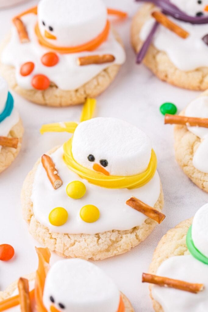Round sugar cookies decorated as melting snowmen with marshmallow heads, icing snow, pretzel arms, candy buttons, and colorful scarf strips.