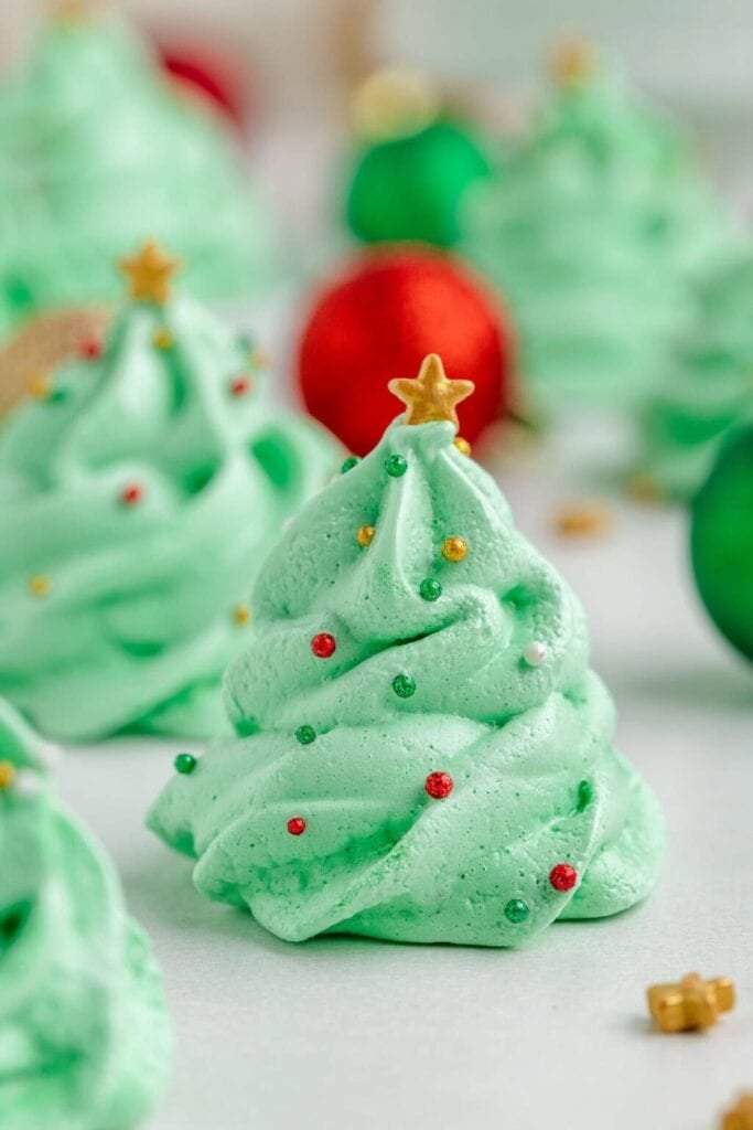 Green meringue cookies shaped like Christmas trees, decorated with colorful sprinkles and golden stars, arranged with blurred festive ornaments in background.