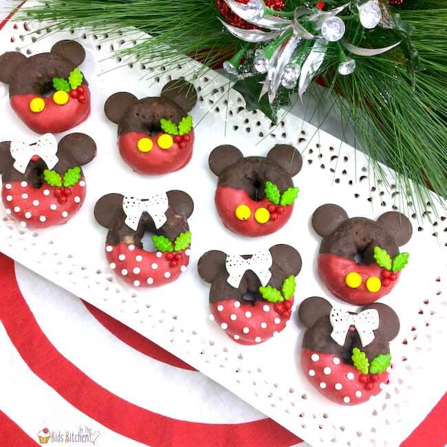 Festive donuts decorated as Mickey and Minnie with chocolate ears, red icing, bows or buttons, and holly details, arranged on tray with holiday greenery and ornaments.