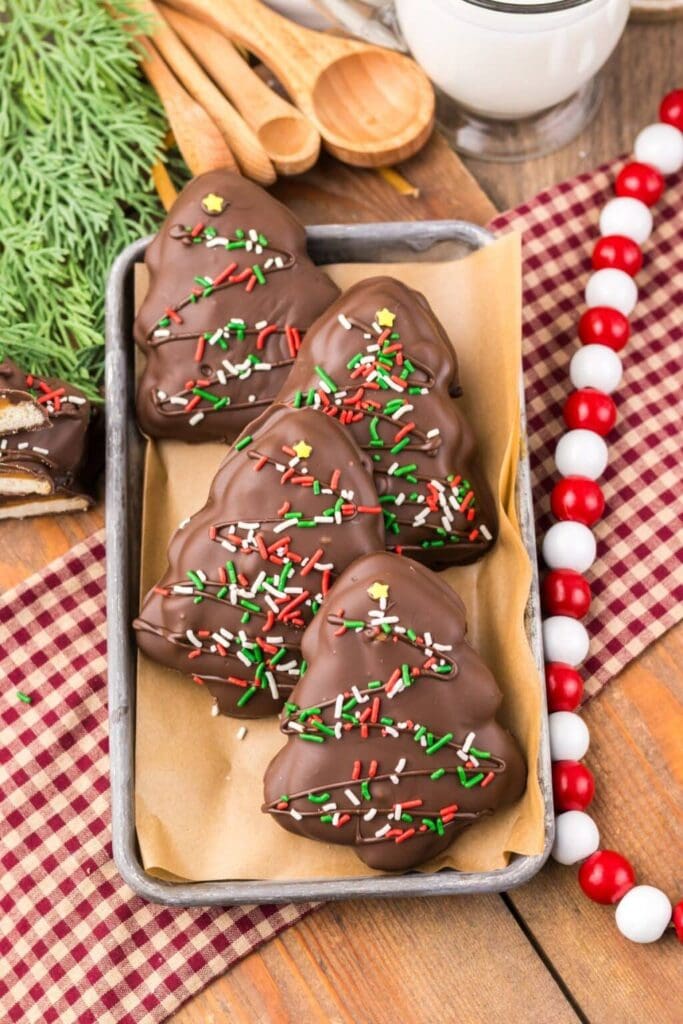 Tray of chocolate Christmas tree treats decorated with red, white, and green sprinkles, topped with yellow stars, set on wooden surface with festive cloth, milk, beads, and pine foliage.