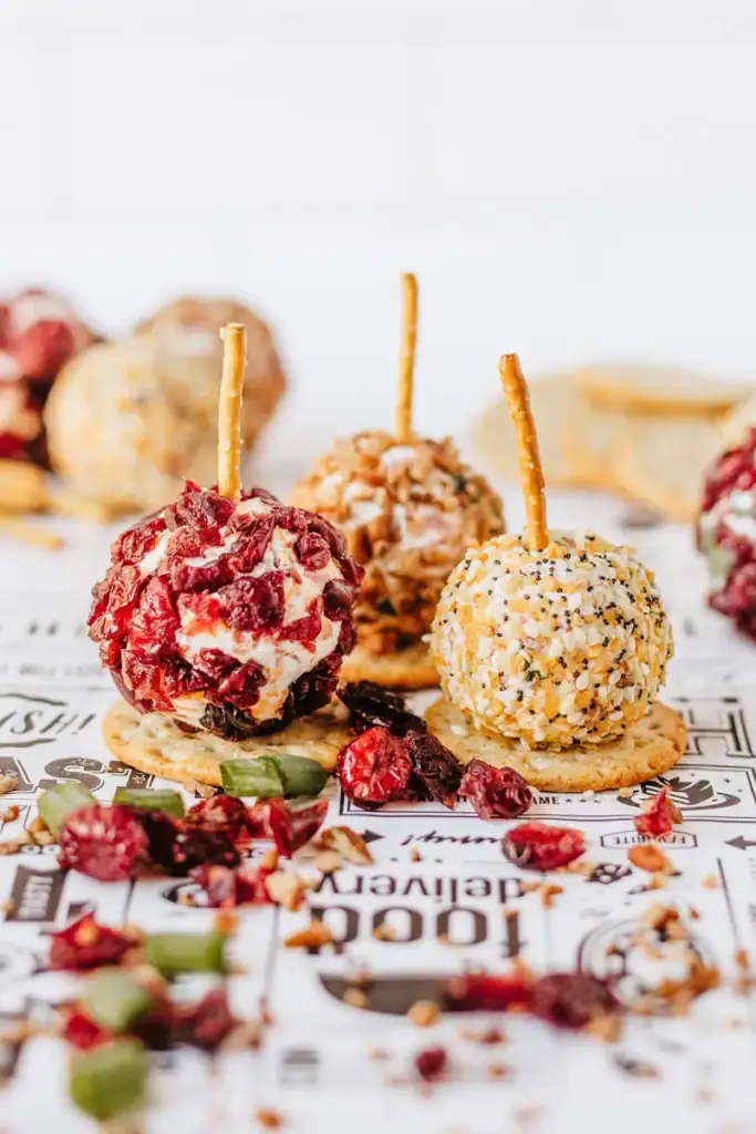 Three mini cheese balls on crackers with pretzel sticks, coated in cranberries, nuts with herbs, and mixed seeds, surrounded by scattered toppings.