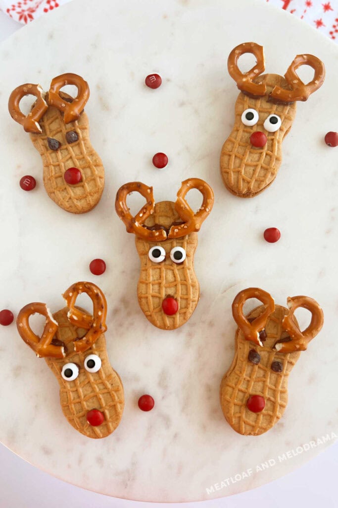 Nutter Butter cookies decorated as reindeer with pretzel antlers, candy eyes, and red noses, arranged on white marble surface with scattered red candies.