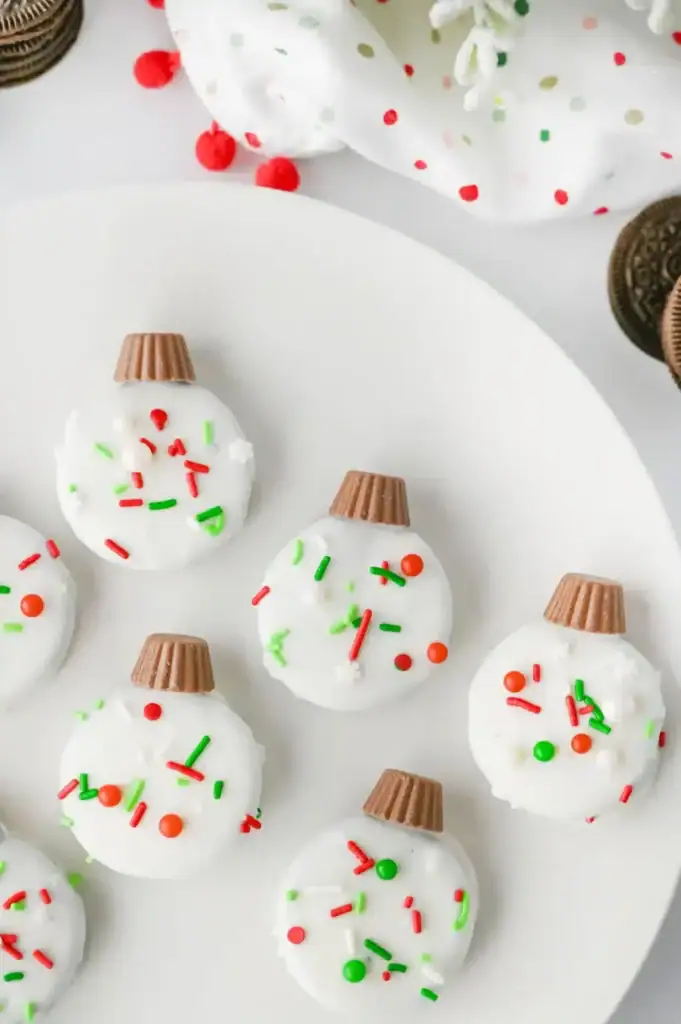 Oreo cookies coated in white chocolate with colorful sprinkles and peanut butter cup tops, arranged on festive plate with polka-dot cloth and red pom-poms.