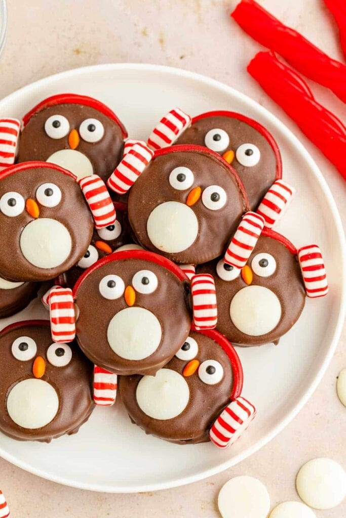 Chocolate cookies decorated as penguins with white candy bellies, candy eyes, orange beaks, and red earmuffs, arranged on white plate with festive candies nearby.