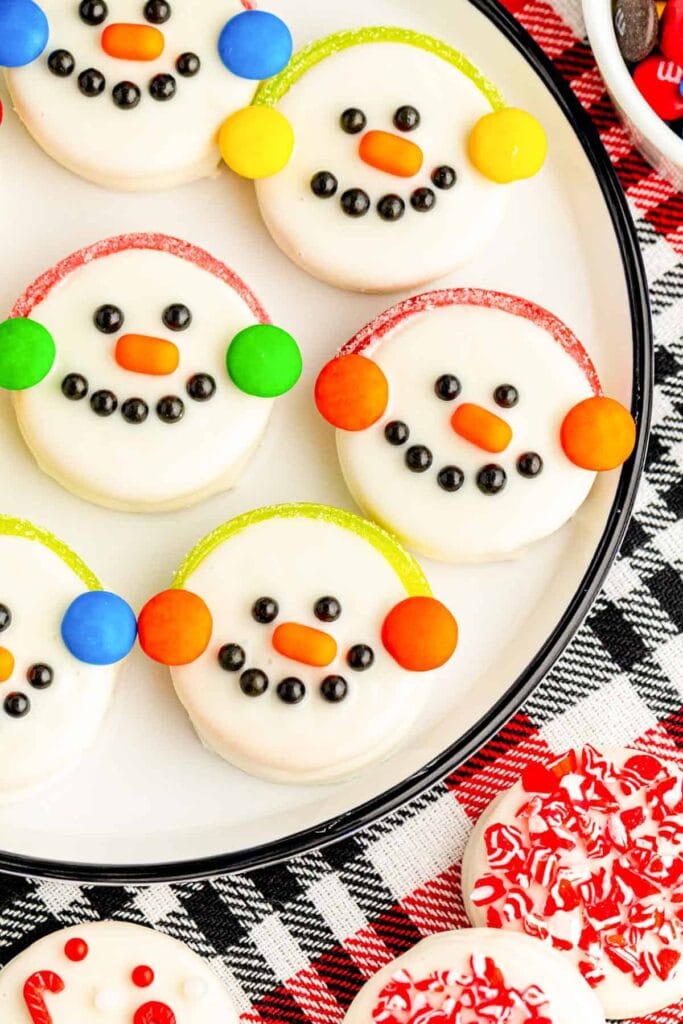 Oreos decorated as snowman faces with icing, candy eyes, orange noses, and colorful earmuff candies, arranged on white plate over plaid cloth.