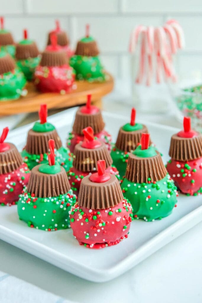 Round red and green candy-coated treats decorated with sprinkles, topped with upside-down peanut butter cups and licorice hooks to resemble Christmas ornaments, arranged on tray with candy canes and sprinkles.