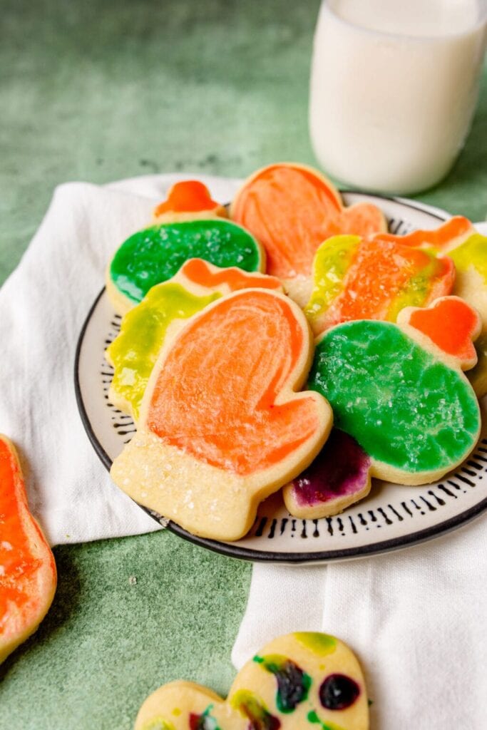 Colorful mitten-shaped sugar cookies frosted in bright orange, green, yellow, and purple with sugar crystals, arranged on plate with milk glass in background.