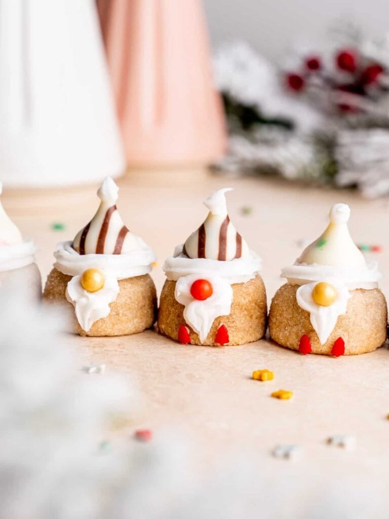Mini desserts decorated as gnomes with frosting beards, striped chocolate or frosting hats, and candy noses, arranged with festive greenery and pastel holiday decor.