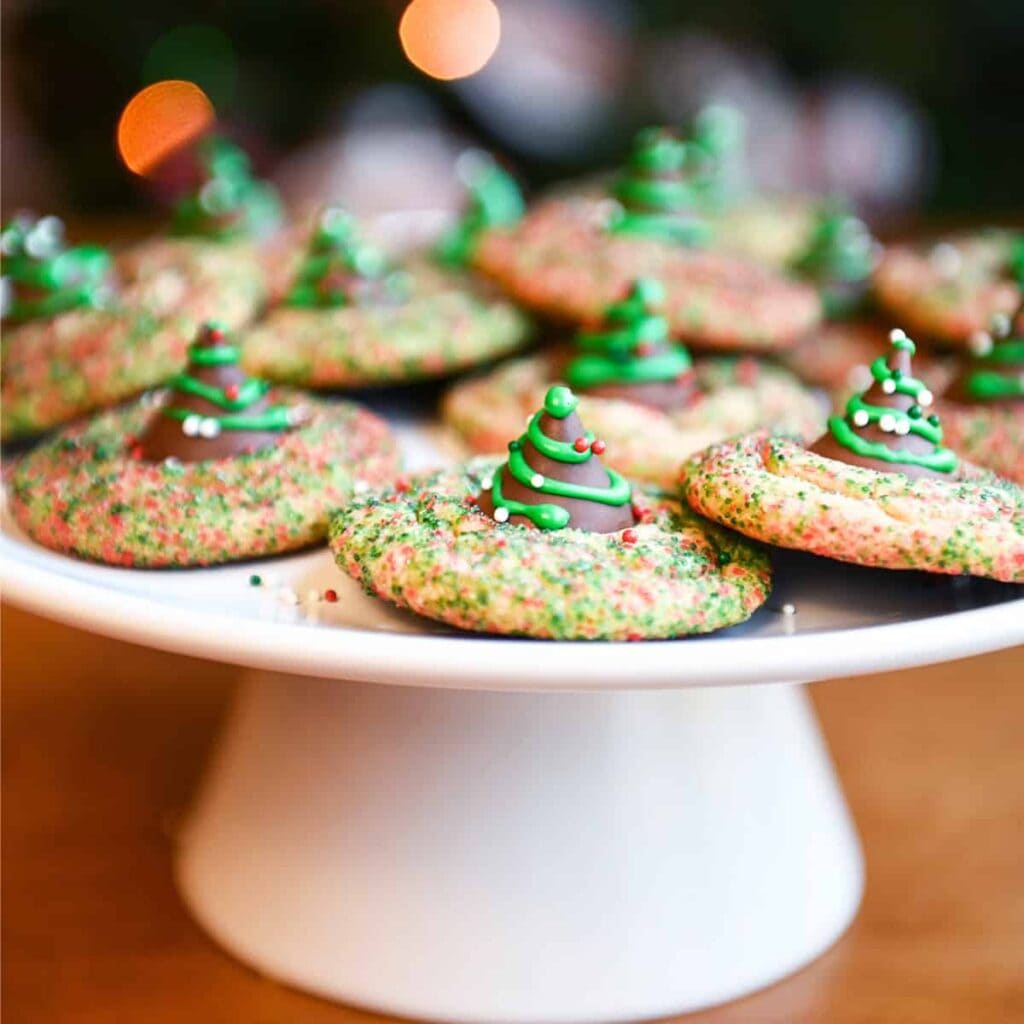 Round cookies topped with chocolate Hershey’s Kiss decorated as green Christmas trees with sprinkles, arranged on white cake stand against warm festive lights.