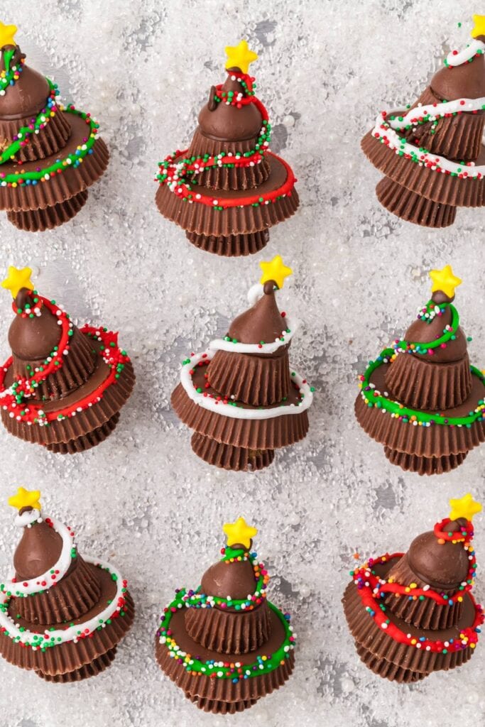 Stacked peanut butter cups topped with chocolate kiss, decorated as Christmas trees with icing, sprinkles, and star candies on snowy white background.