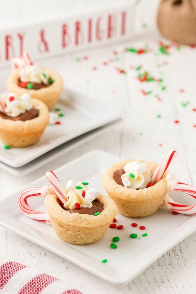 Cookie cups filled with chocolate, topped with whipped cream, marshmallows, sprinkles, and mini candy canes, arranged on plates with festive background.