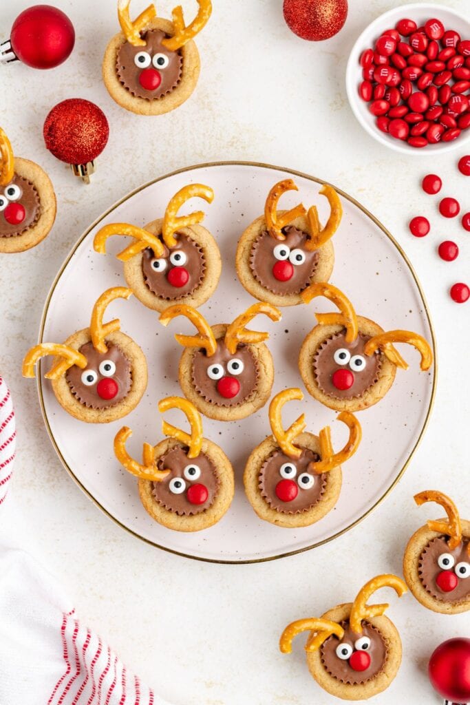 Round cookies decorated as reindeer faces with candy eyes, red noses, and pretzel antlers, arranged on white plate with gold rim among red ornaments and M&M bowl.
