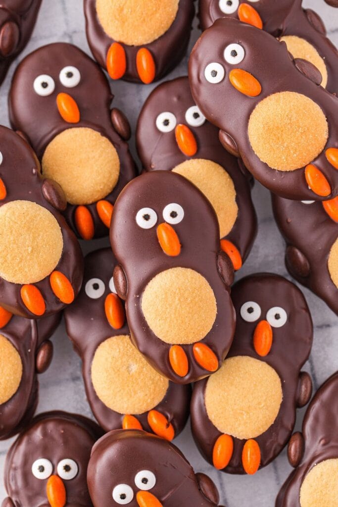 Chocolate-coated cookies decorated as penguins with tan bellies, candy eyes, orange beaks, and feet, arranged closely on light surface.