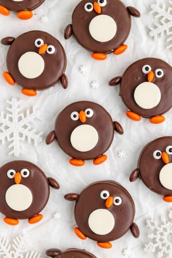 Round chocolate cookies decorated as penguins with candy eyes, orange beaks and feet, and oval wings, arranged on white surface with snowflake sprinkles.