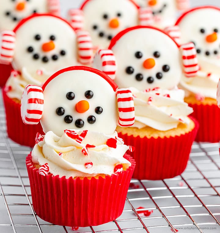 Red cupcake wrappers with frosted snowman faces, candy earmuffs, and peppermint sprinkles, arranged on cooling rack.