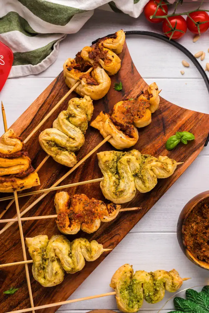 Wooden board with skewered puff pastry twists topped with pesto and tomato spreads, surrounded by basil, cherry tomatoes, pine nuts, and sauce bowl.