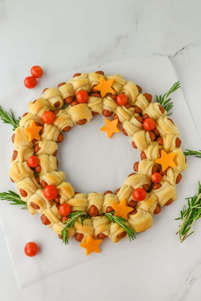 Wreath-shaped arrangement of golden crescent-wrapped sausages, decorated with rosemary sprigs, cherry tomatoes, and star-shaped cheese on a white surface.