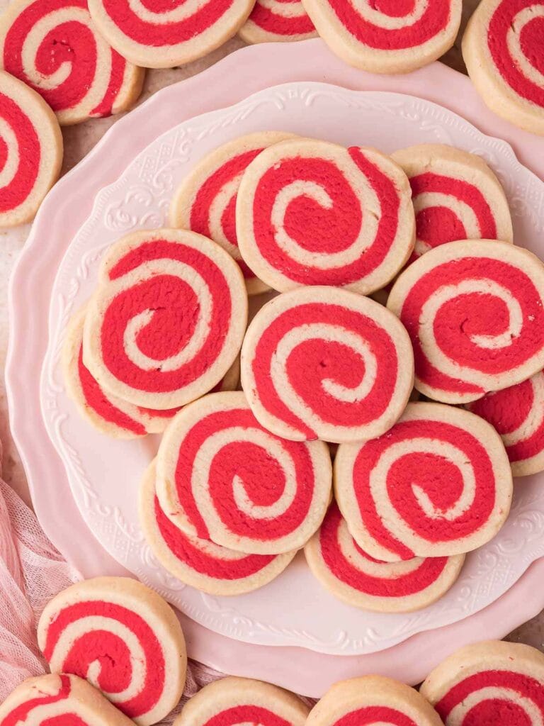 Red and white pinwheel sugar cookies arranged on decorative white plate over soft pink surface with fabric nearby.
