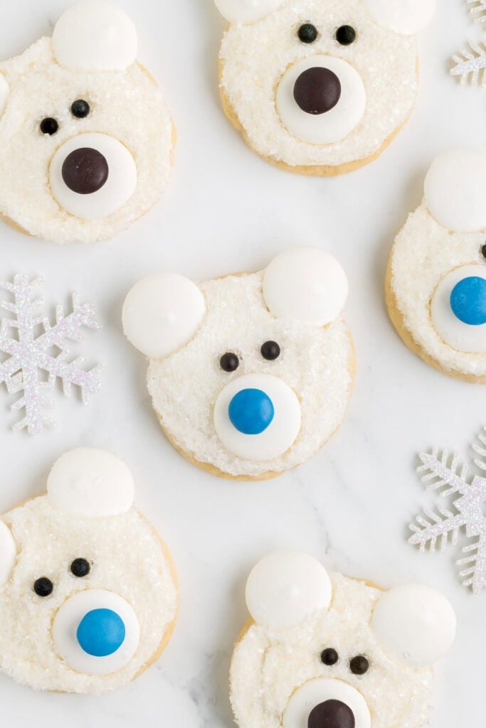 Round cookies decorated as polar bear faces with white frosting, sugar crystal texture, candy ears, dark eyes, and colorful noses, arranged on white surface with glittery snowflake accents.