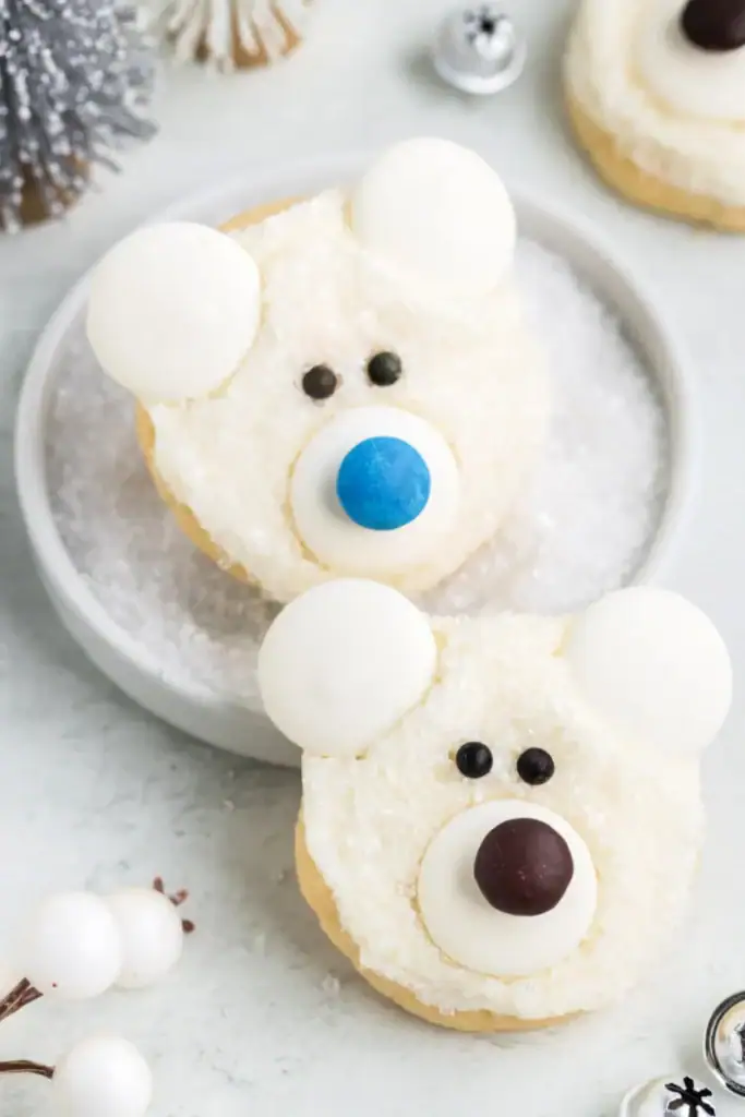 Round cookies decorated as polar bear faces with white frosting, sugar crystal fur, candy ears, dark eyes, and colorful noses, set among snowy decorations and silver bells.