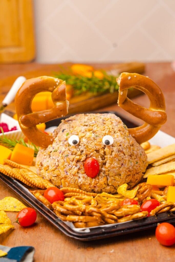 Cheese ball coated in nuts decorated as a reindeer with pretzel antlers, candy eyes, and tomato nose, surrounded by crackers, pretzels, cheese cubes, and cherry tomatoes on a tray.