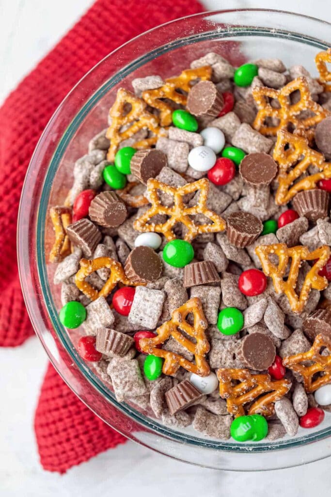 Glass bowl of holiday snack mix with chocolate cereal, peanut butter cups, candy-coated chocolates, and star pretzels on red cloth.