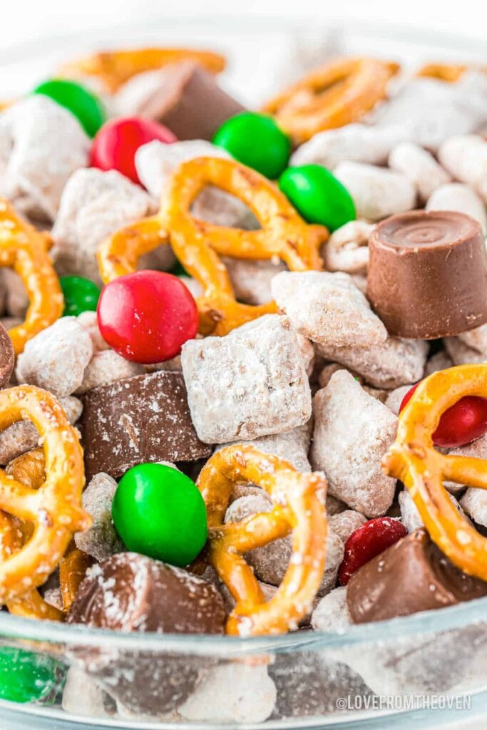 Glass bowl of holiday snack mix with pretzels, red and green candies, chocolate caramels, and powdered cereal pieces.