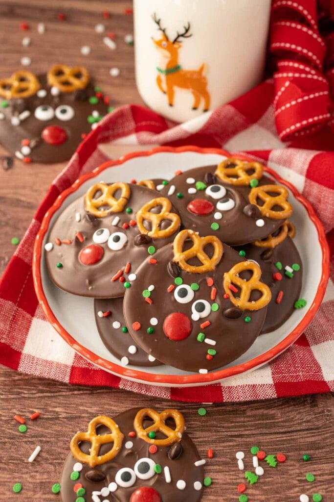 Plate of round chocolate reindeer treats with pretzel antlers, candy eyes, red noses, and holiday sprinkles, set on red-rimmed plate with festive cloth and reindeer cup nearby.