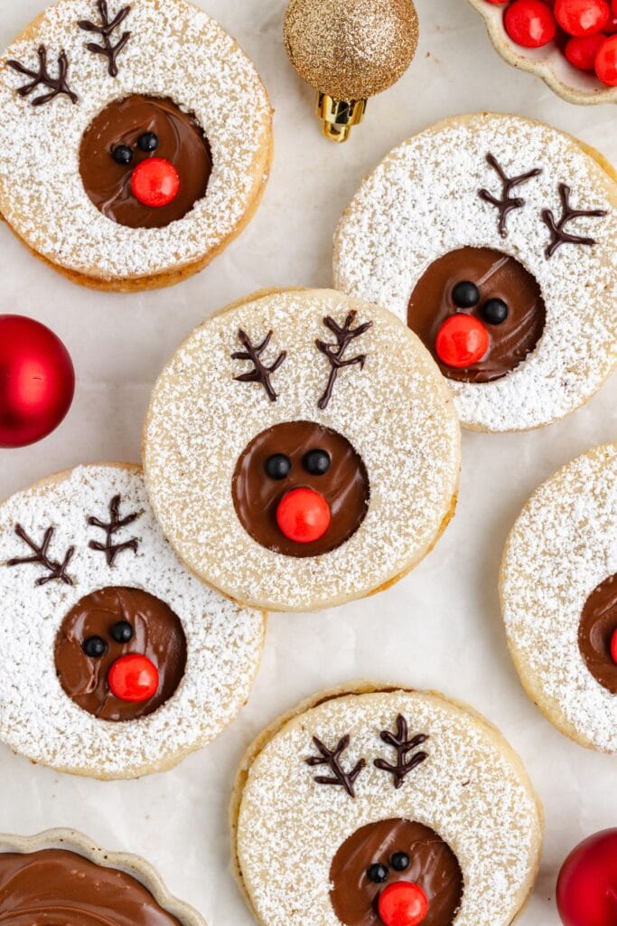 Round cookies with chocolate centers decorated as reindeer faces with candy eyes, red noses, powdered sugar, and piped chocolate antlers, surrounded by red and gold ornaments.
