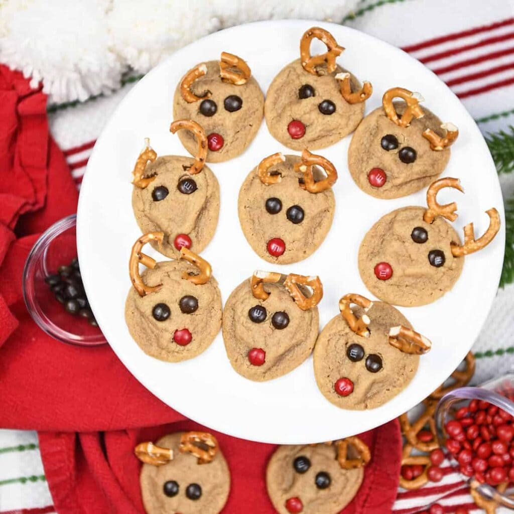 Round cookies decorated as reindeer with pretzel antlers, candy eyes, and red noses, arranged neatly on white plate with festive red cloth.