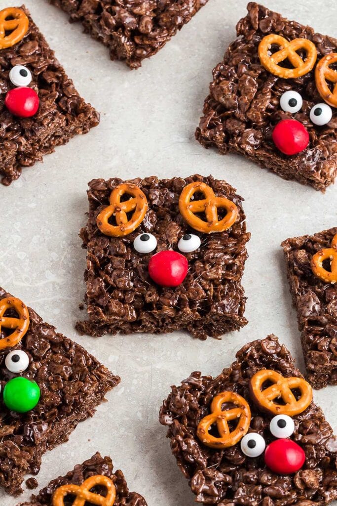 Chocolate Rice Krispies squares decorated as reindeer with pretzel antlers, candy eyes, and red or green noses, arranged on light surface.