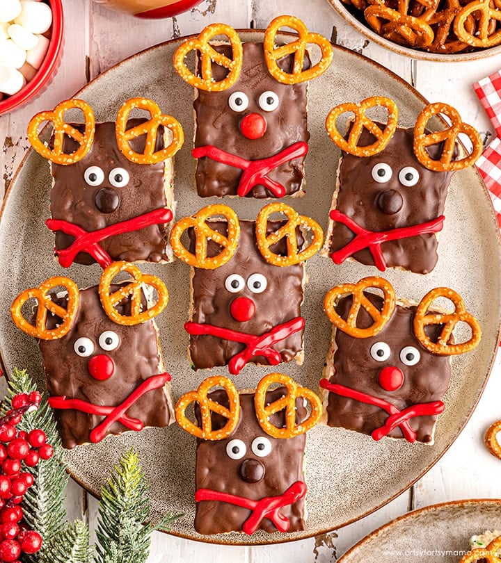 Plate of rectangular chocolate reindeer treats with pretzel antlers, candy eyes, red or brown noses, and licorice scarves, arranged on rustic wood with festive greenery and berries.
