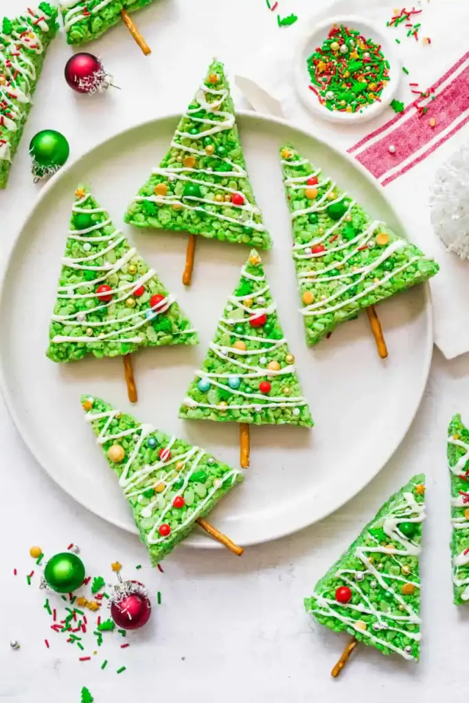 Green Rice Krispies Christmas tree treats with icing drizzles, candy ornaments, and pretzel trunks, arranged on white plate with festive sprinkles and ornaments.