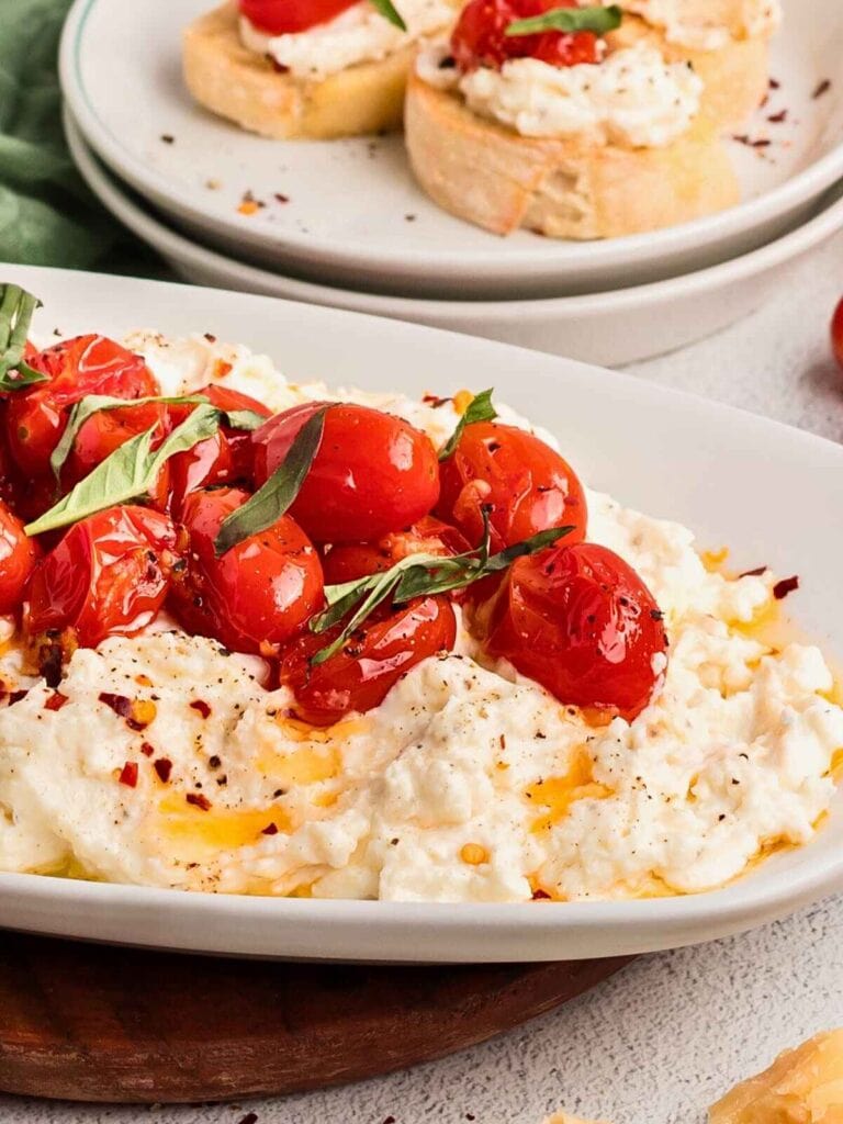 Creamy ricotta topped with roasted cherry tomatoes, basil, olive oil, and pepper, served with toasted bread slices in background.