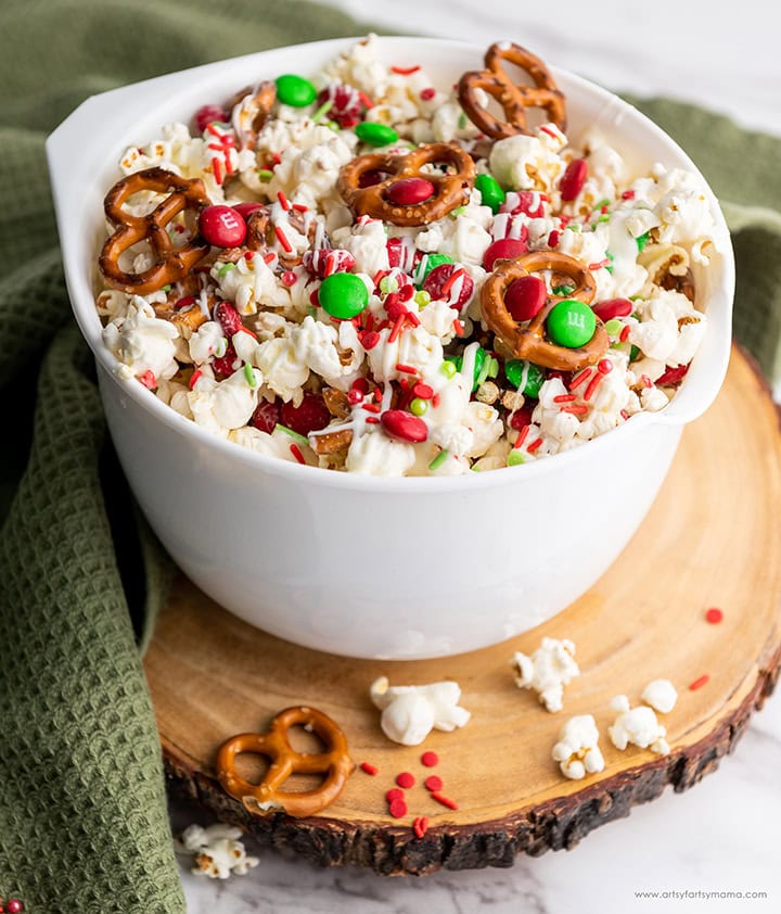 White bowl of popcorn snack mix with pretzels, red and green candies, sprinkles, and white chocolate drizzle, set on wooden board with green cloth.