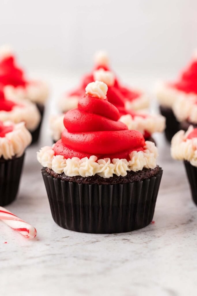 Chocolate cupcake in black liner topped with red frosting swirl shaped like Santa hat, finished with white dollop and trim, with candy cane nearby.