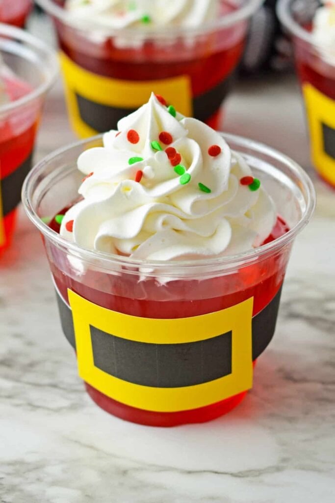 Clear cup of red gelatin topped with whipped cream and sprinkles, wrapped in black paper band with yellow buckle for Santa design, set among similar festive cups.