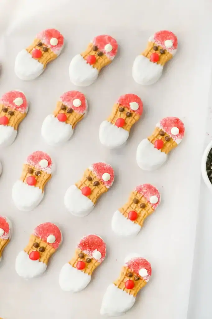 Peanut butter sandwich cookies decorated as Santa faces with red sugar hats, white icing beards, chocolate chip eyes, and red candy noses, arranged neatly on white surface.